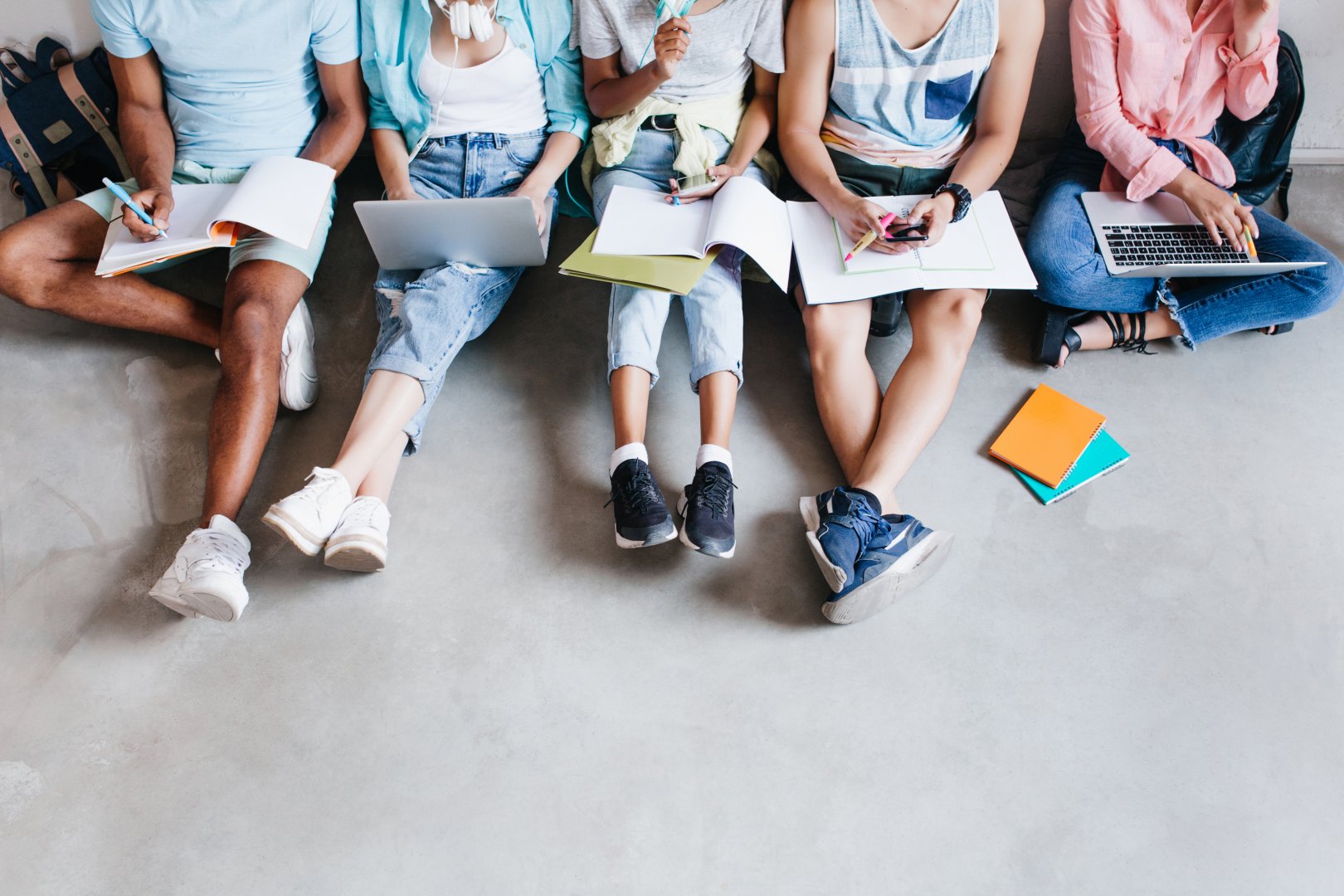 Overhead portrait of young people with laptops and smartphones, sitting together on the floor. Students writing lectures holding textbooks on their knees..